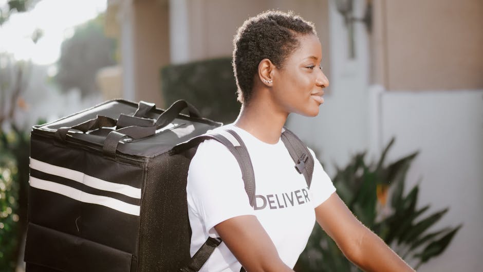 A young woman with short, curly hair and dark skin, dressed in a white T-shirt with 'DELIVER' printed on it, is carrying a large black delivery bag with white stripes on her back using adjustable shou