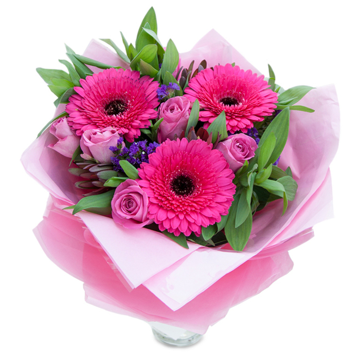I see a bouquet composed with the kind of confident colour pairing that lifts a room: three fuchsia Gerberas dominate the scene, their saturated petals arranged in precise rays around vivid, dark centres that give them presence and personality. Nestled among them are delicate roses in a dusty lavender-pink, their buds still holding form so they feel fresh and resolutely new. Interspersed are little clusters of purple statice which bring a wildflower sensibility and a pleasing textural contrast to the rounded forms of the roses and gerberas. Supporting foliage-broad glossy leaves that read as salal, alongside softer eucalyptus sprigs-creates a lush green backdrop that both grounds and elevates the pink tones. The bouquet is lovingly wrapped: a soft, matte pale pink tissue inner layer cushions the stems while an outer, translucent fuchsia-pink cellophane with a slight crinkle lends a gentle sheen and adds a crafted finish. The lower edge reveals the top rim of a clear glass vase, a promise of an immediate, effortless display in a hallway or kitchen. Imagine receiving this after a walk past the RAF Museum or a quick stop at Colindale tube: its scent would be light and rosy, its textures velvety and crisp, and its presence would make a birthday table, Mother's Day breakfast, or anniversary evening feel warmly celebrated. Hand-tied in spirit, the arrangement speaks of thoughtful care and local florist craft tailored to Colindale's everyday moments.